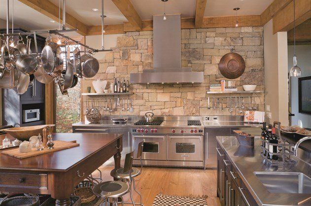 A rustic kitchen featuring a large stone wall, stainless steel appliances, a wooden island, and hanging cookware.