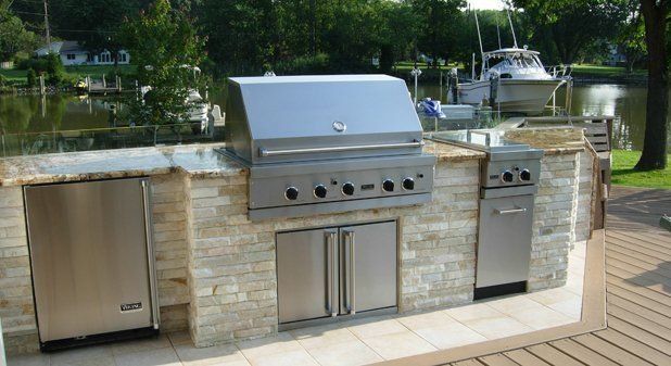 Outdoor stone kitchen with a stainless steel grill, cabinets, and a refrigerator on a deck overlooking a river and boat.
