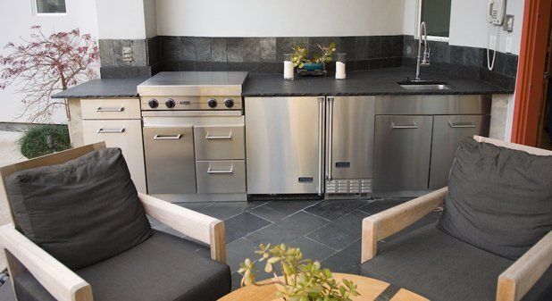 An outdoor kitchen with stainless steel appliances and a dark stone countertop, flanked by two chairs on a slate patio.