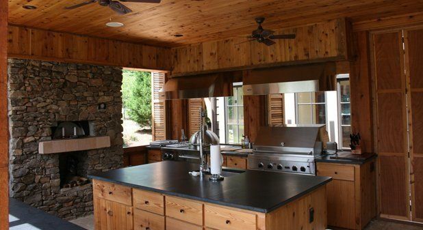 A rustic outdoor kitchen with a stone fireplace, wood paneling, a dark countertop island, and built-in stainless grill.