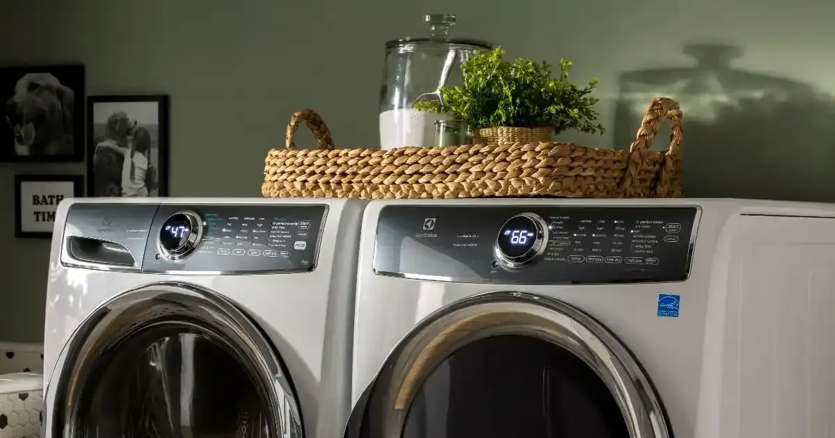 A white side-by-side washer and dryer set with a woven tray, a glass jar, and a small plant on top.