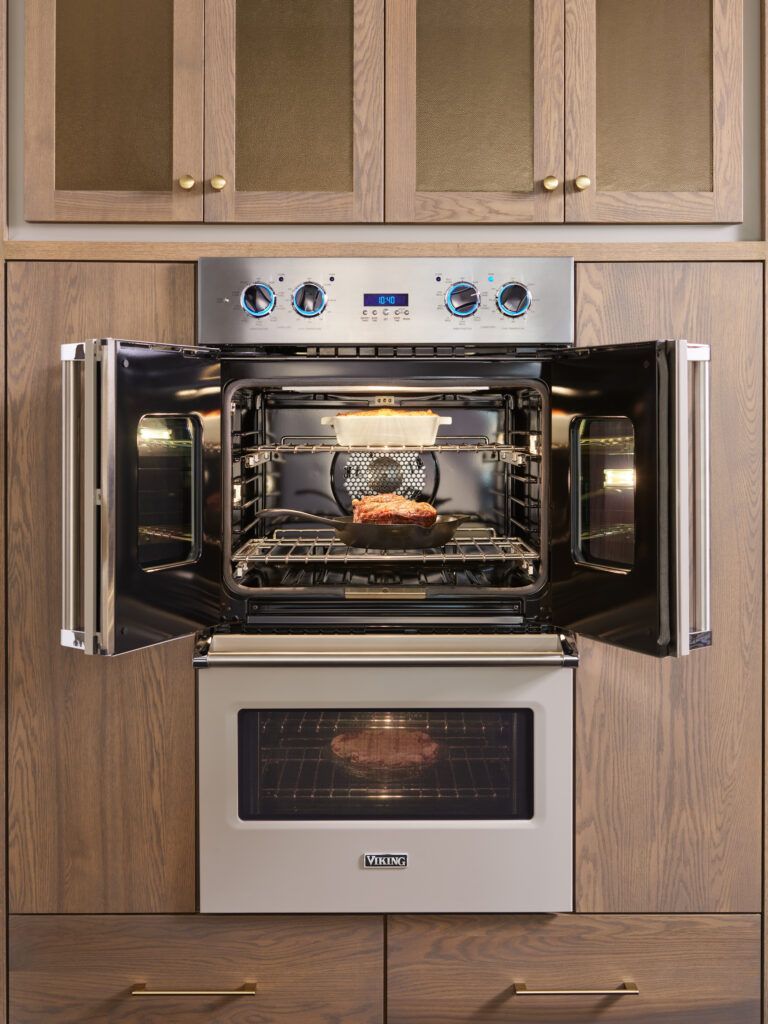 A stainless steel double wall oven set in wood cabinetry, with the top unit's French doors open to reveal food inside.