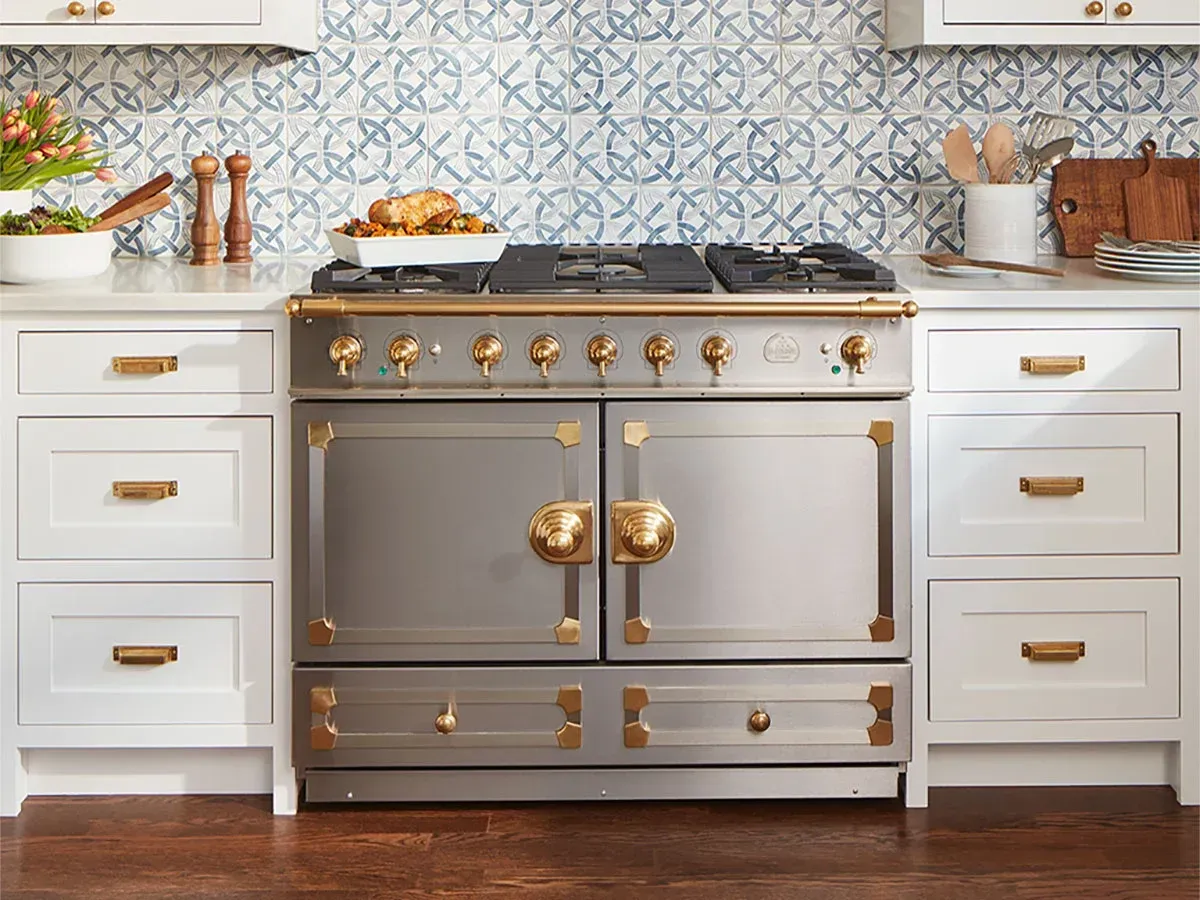 A stainless steel range stove with brass accents centered between white cabinets and a blue patterned tile backsplash.