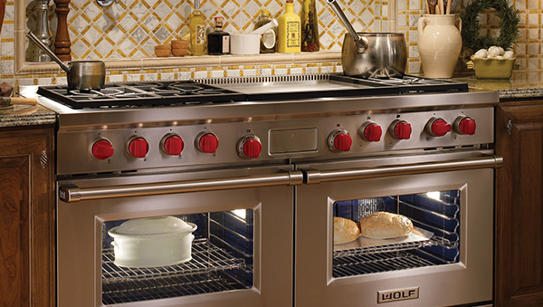 A stainless steel Wolf dual-oven range with iconic red knobs, set into a wooden kitchen cabinet with a tiled backsplash.