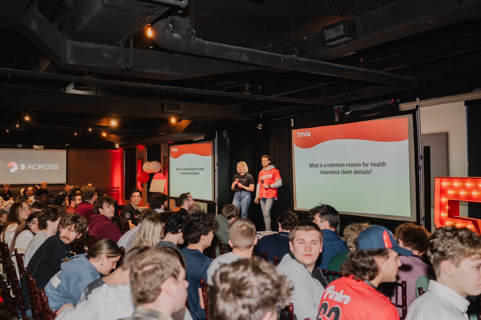 People watch two people speaking on stage with projected text in an event space.