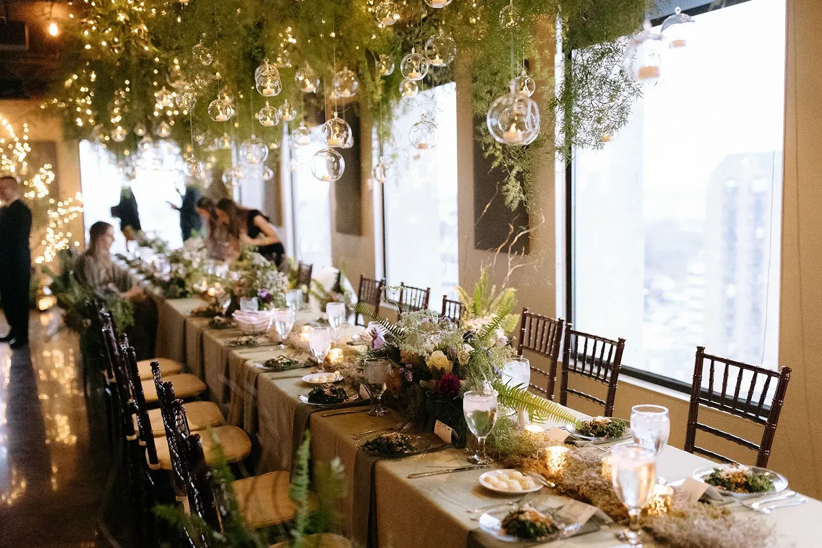 Long dining table set for event, decorated with flowers and hanging orbs, near window.