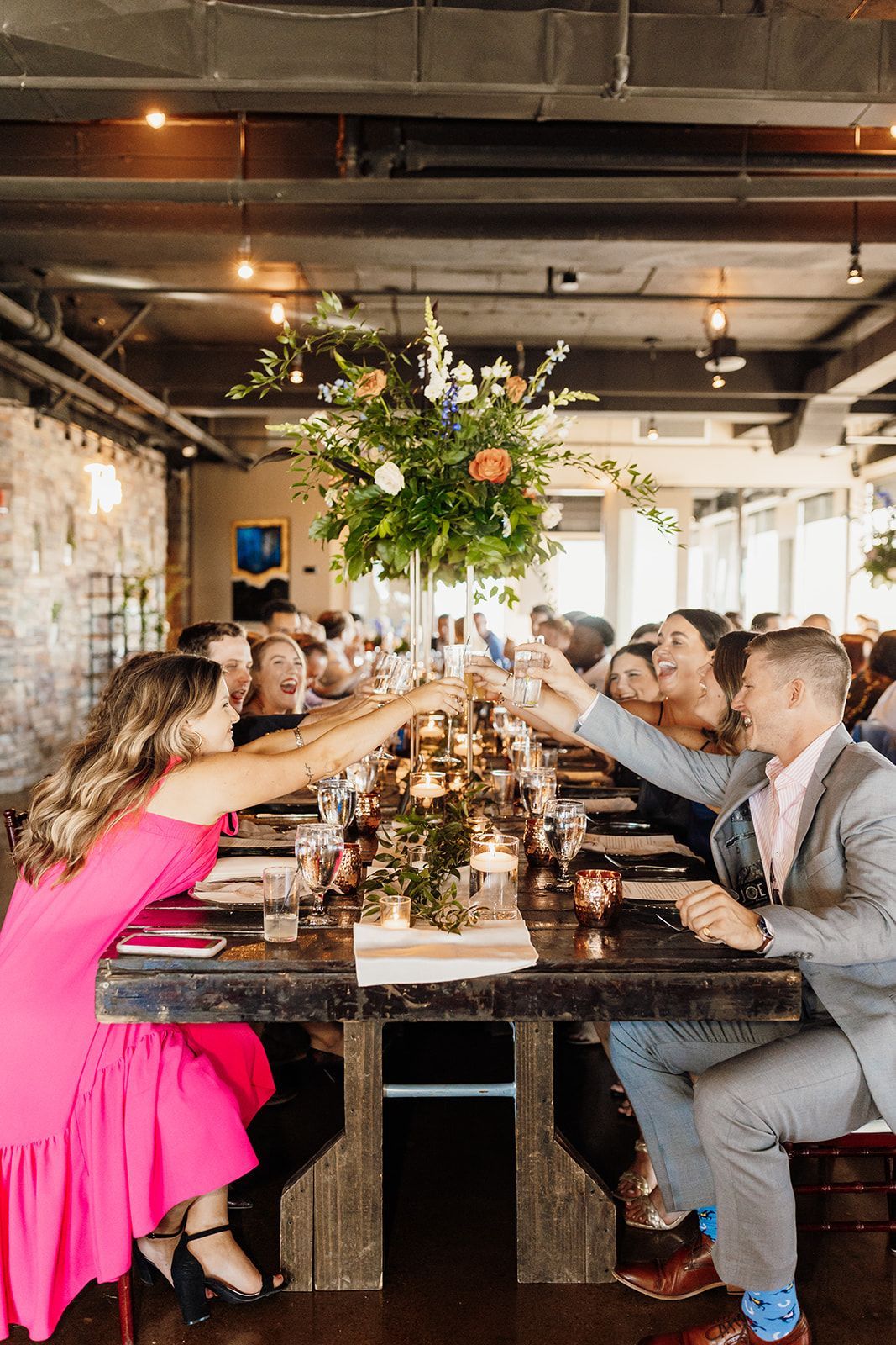 People toasting at a long wooden table set for a celebration, with a large floral centerpiece and a woman in pink.