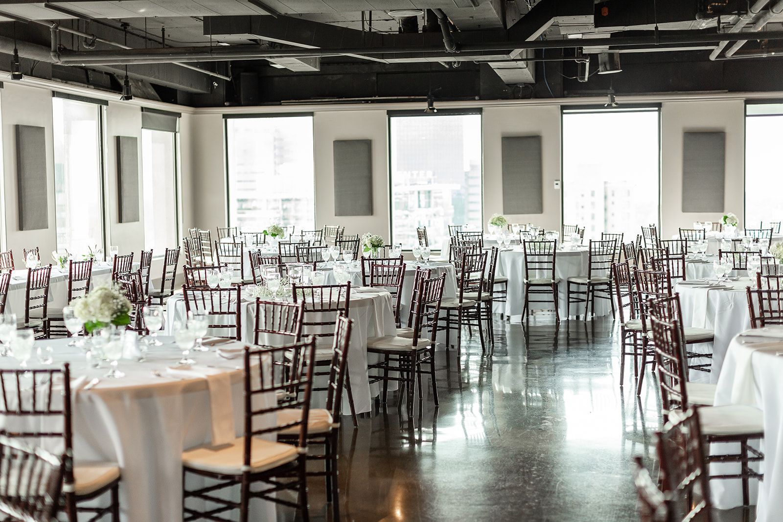 An elegant event space with round tables covered in white linens, brown chairs, and large windows.