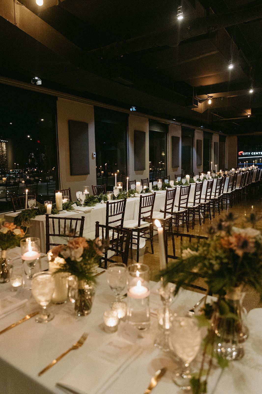 Long wedding reception tables set with candles, flowers, and place settings; windows with a city view in the background.