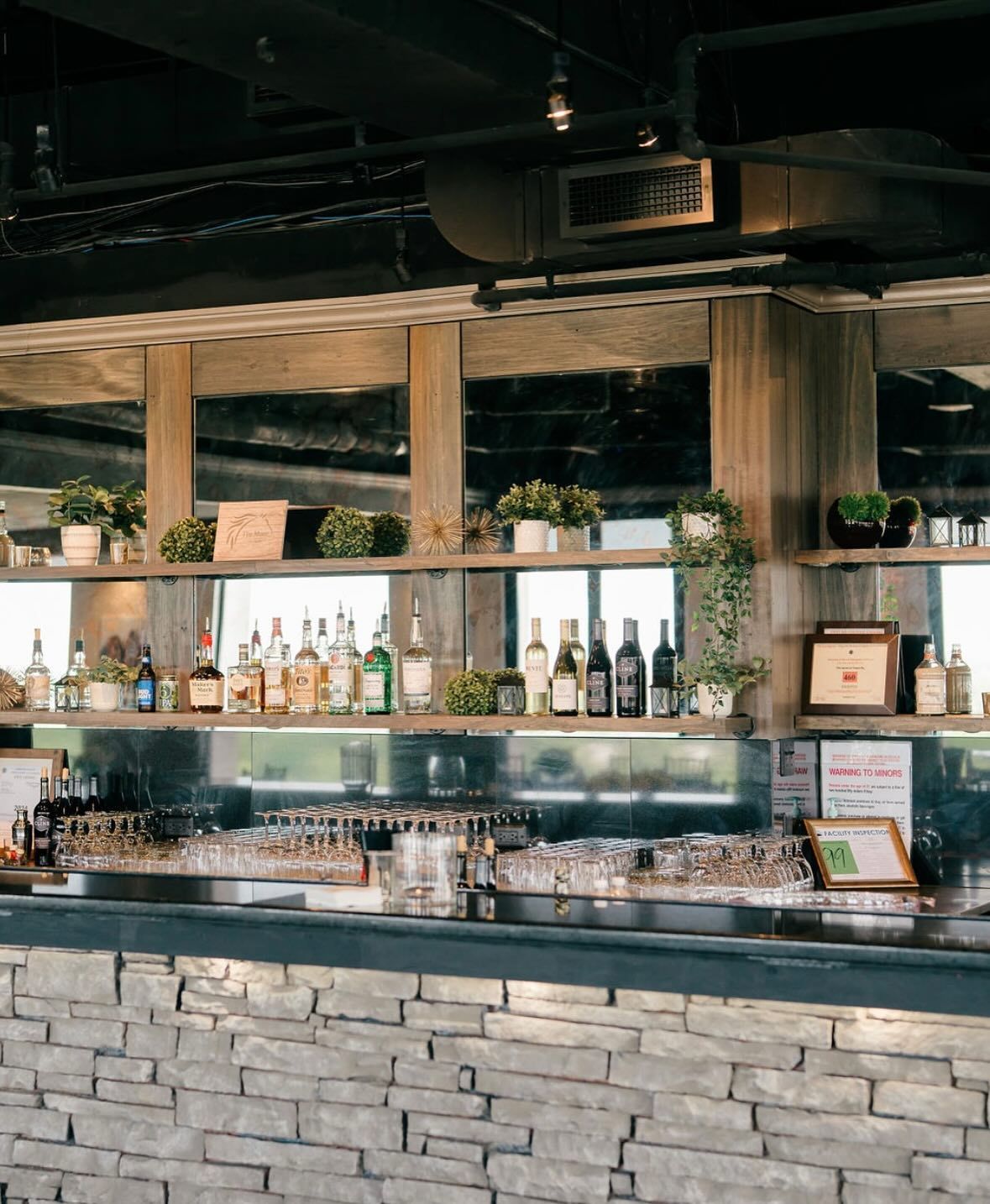 Stone-faced bar with liquor bottles, glassware, and potted plants. Mirrors and shelves are behind the bar.
