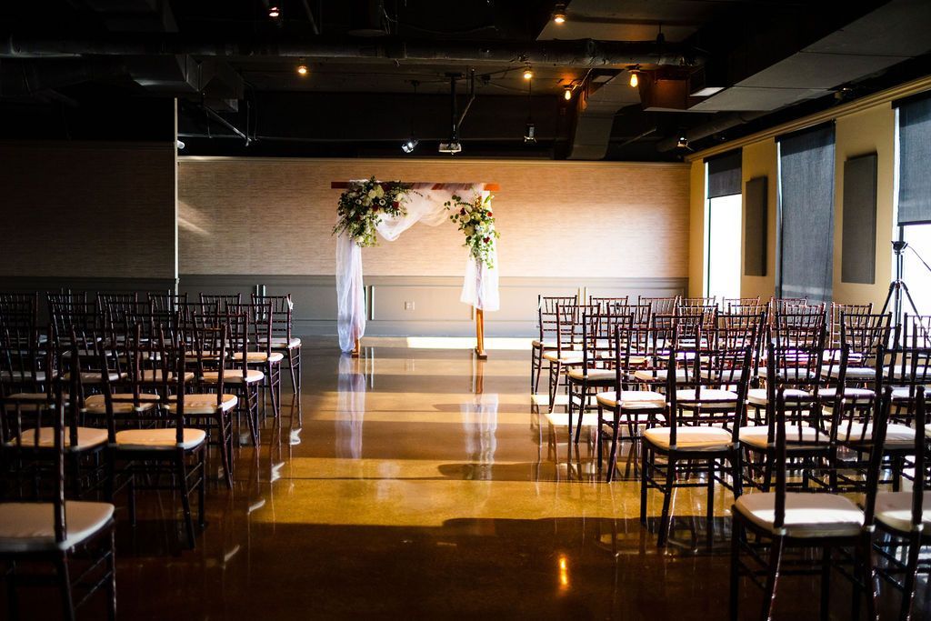 Empty event space set up for a wedding ceremony, with rows of chairs facing an arch decorated with flowers and fabric.