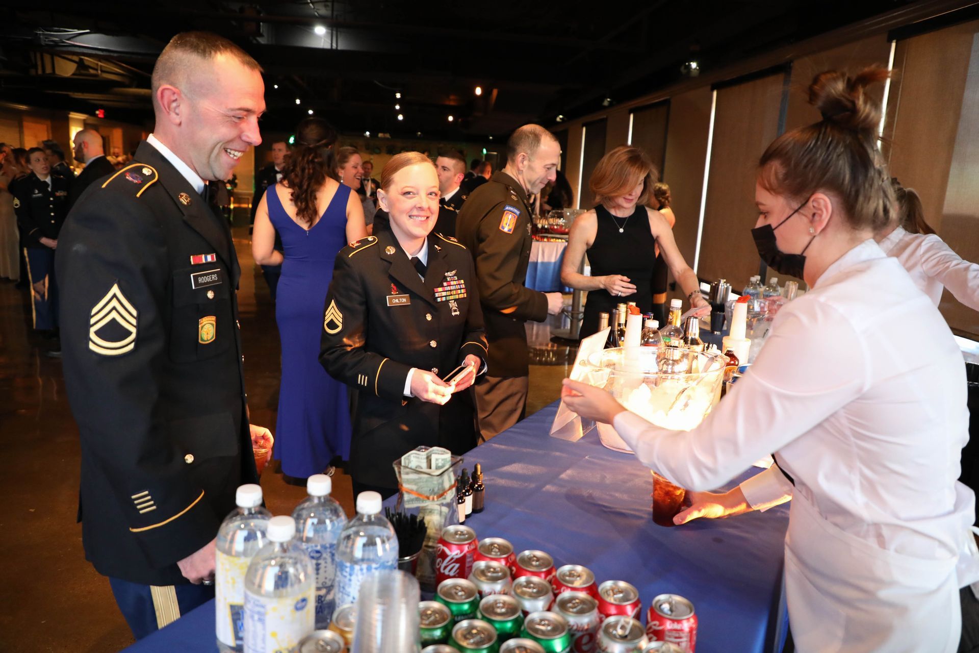 Soldiers in formal attire at a bar, smiling at a server with drinks. People in background at a formal event.