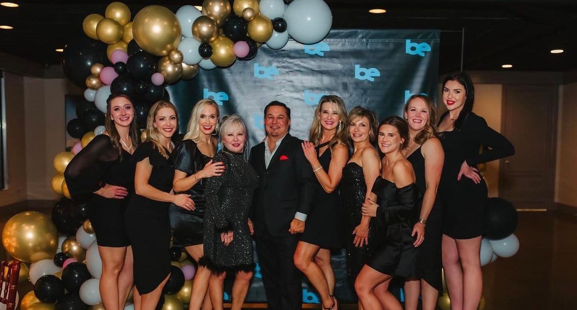 Group of people posing in front of a backdrop with balloons. Most are women wearing black dresses, smiling, and posing.