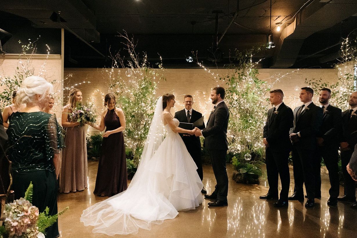 Wedding ceremony: Bride and groom stand before a minister in a room with guests seated. Large windows and greenery frame the scene.