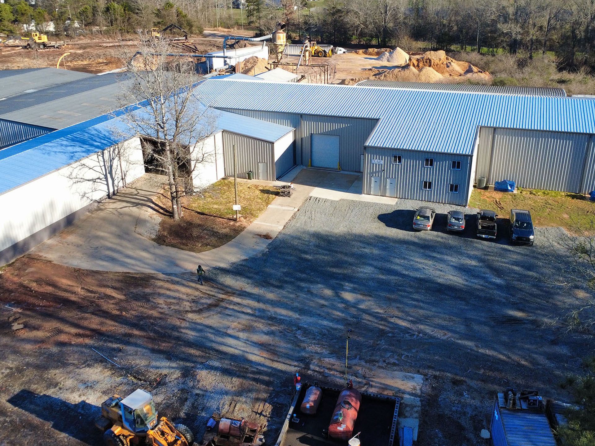 Aerial view of a light-blue industrial building with a paved lot, parked cars, and surrounding trees.