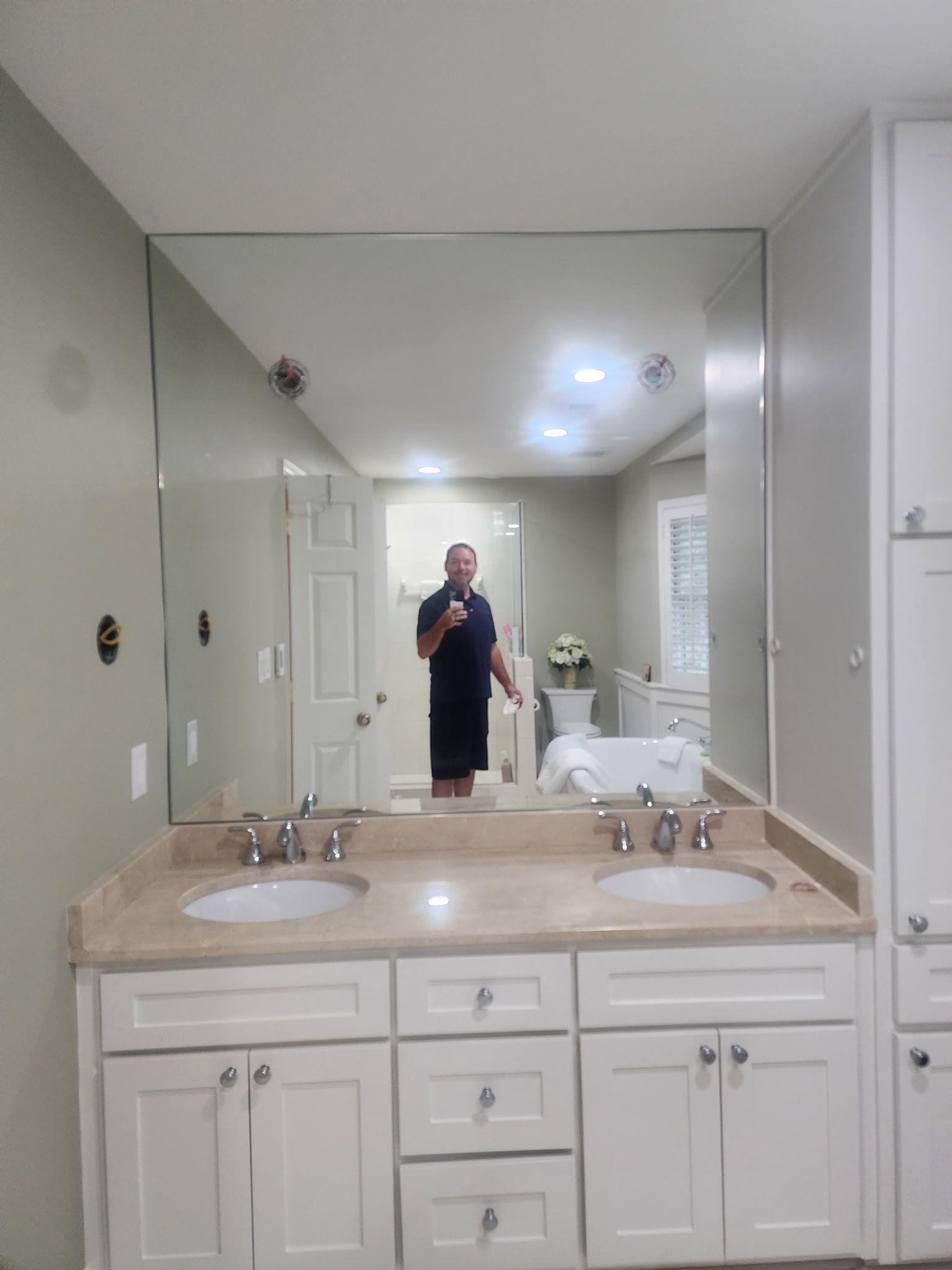 Bathroom with large mirror reflecting a person, double sinks, white cabinets, and beige countertop.