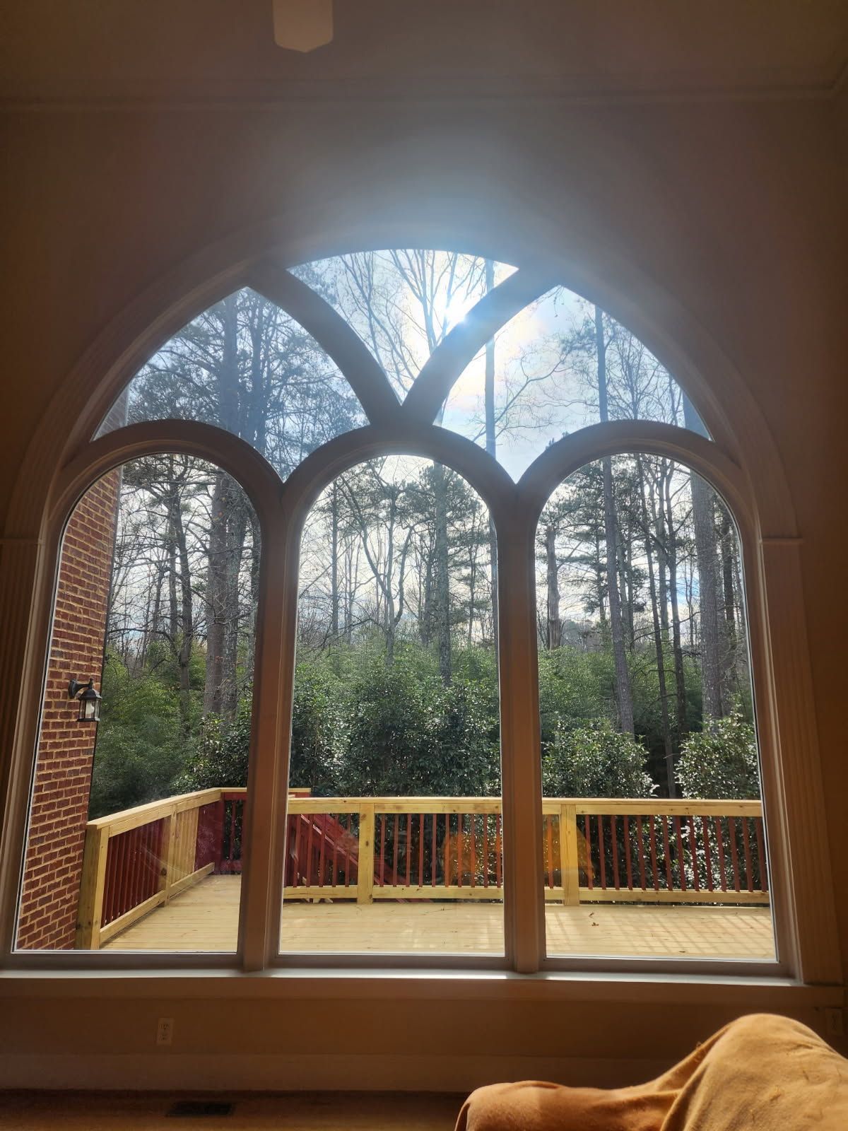 Arched window with three vertical sections, overlooking a wooden deck and trees. Bright sunlight shines through the window.