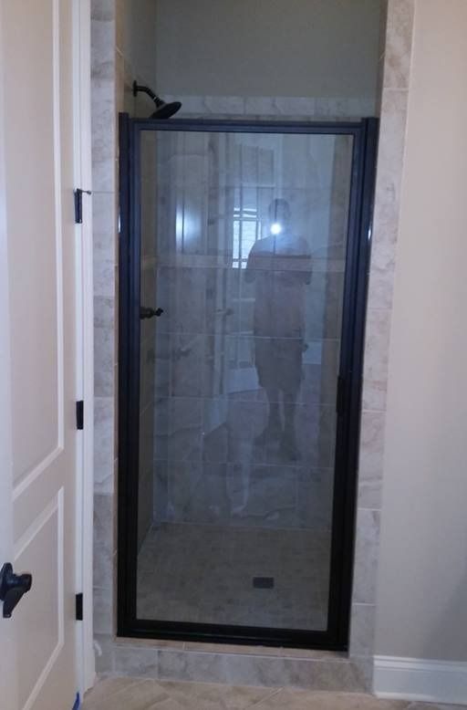 Black-framed shower door with glass. A person is reflected in the glass. Beige tile and a black showerhead are visible.