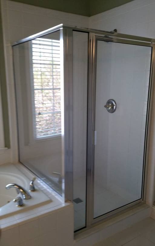 Corner shower with glass door and side panel next to a bathtub; window with blinds in background.