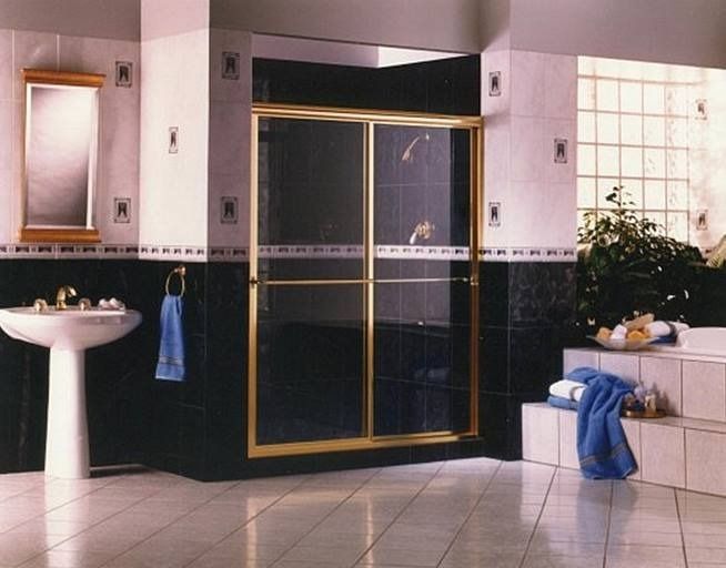 Bathroom with gold-framed glass shower doors, white pedestal sink, black tile, and a bathtub with a plant.