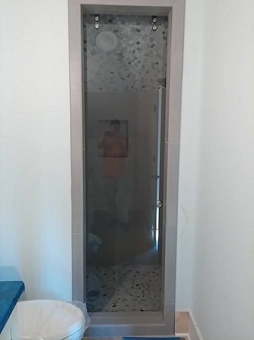 Narrow shower stall with a glass door, framed in gray, built into a bathroom wall with a stone tile floor.