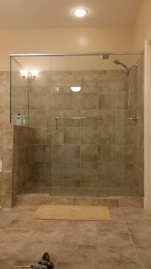 A tiled shower with a glass door, shower head, and a tan bath mat in a bathroom.