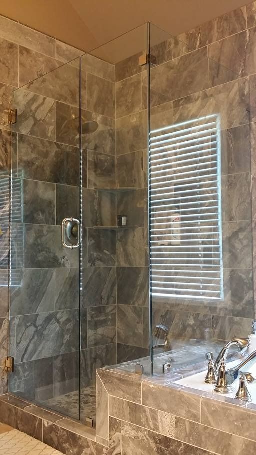 Shower with glass doors and grey tile walls, next to a window with blinds.