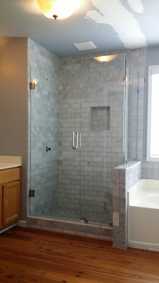 Glass-enclosed shower with gray tiled walls and a wooden floor. Bathroom with a vanity and bathtub visible.