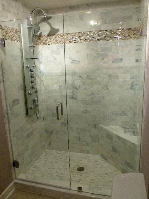 Glass-enclosed shower with white marble tile and a pebble accent. Includes a bench and multiple shower heads.