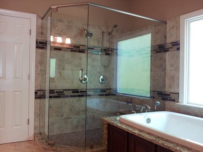 Glass-enclosed shower in a bathroom with a window, tub, and tan tile walls.