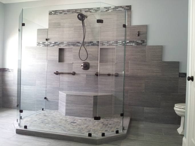 Modern bathroom with large glass shower enclosure; gray tiled walls, shower bench, and pebble floor.