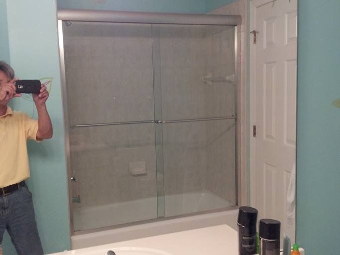 Person taking a photo in a bathroom mirror. Shower with glass doors. Light blue walls and white door visible.