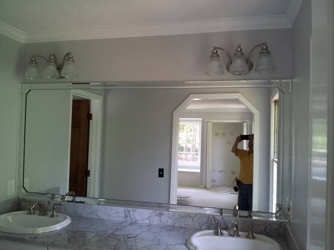 Bathroom with large mirror, two sinks, and overhead lights. A person is visible, reflected in the mirror, taking a photo.