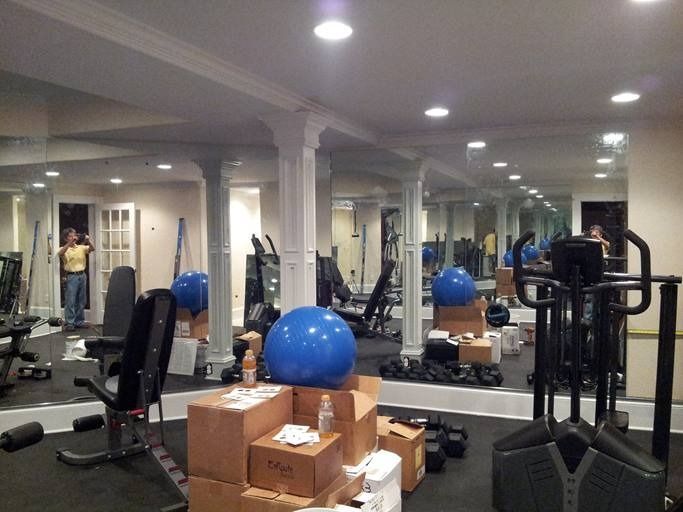 Gym with workout equipment, mirrors, and a person standing in doorway. Boxes and a blue exercise ball are in the middle of the room.