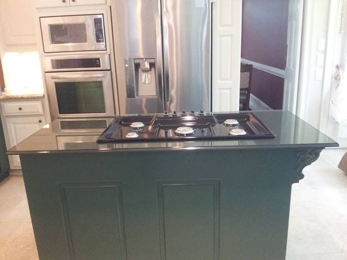 Green kitchen island with a stovetop in front of a stainless steel refrigerator and oven.