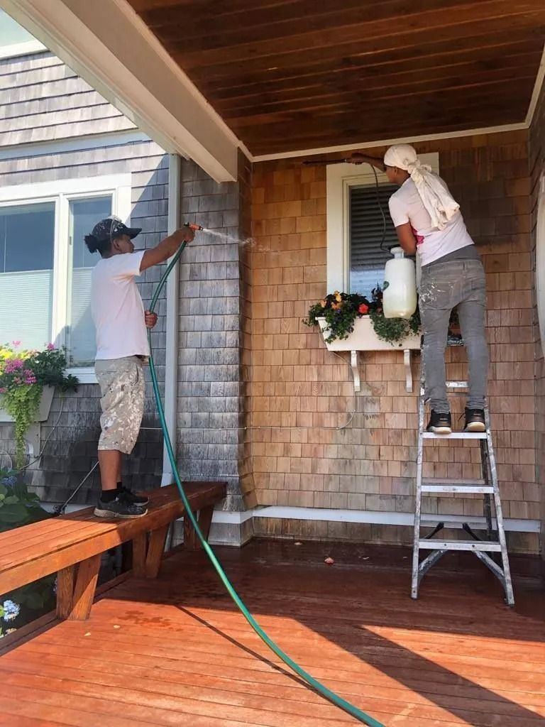 Two people cleaning a house exterior: one sprays with a hose, the other on a ladder by a window.