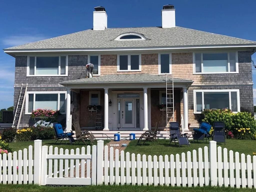 House exterior being painted, with a worker on a ladder. White picket fence in front.