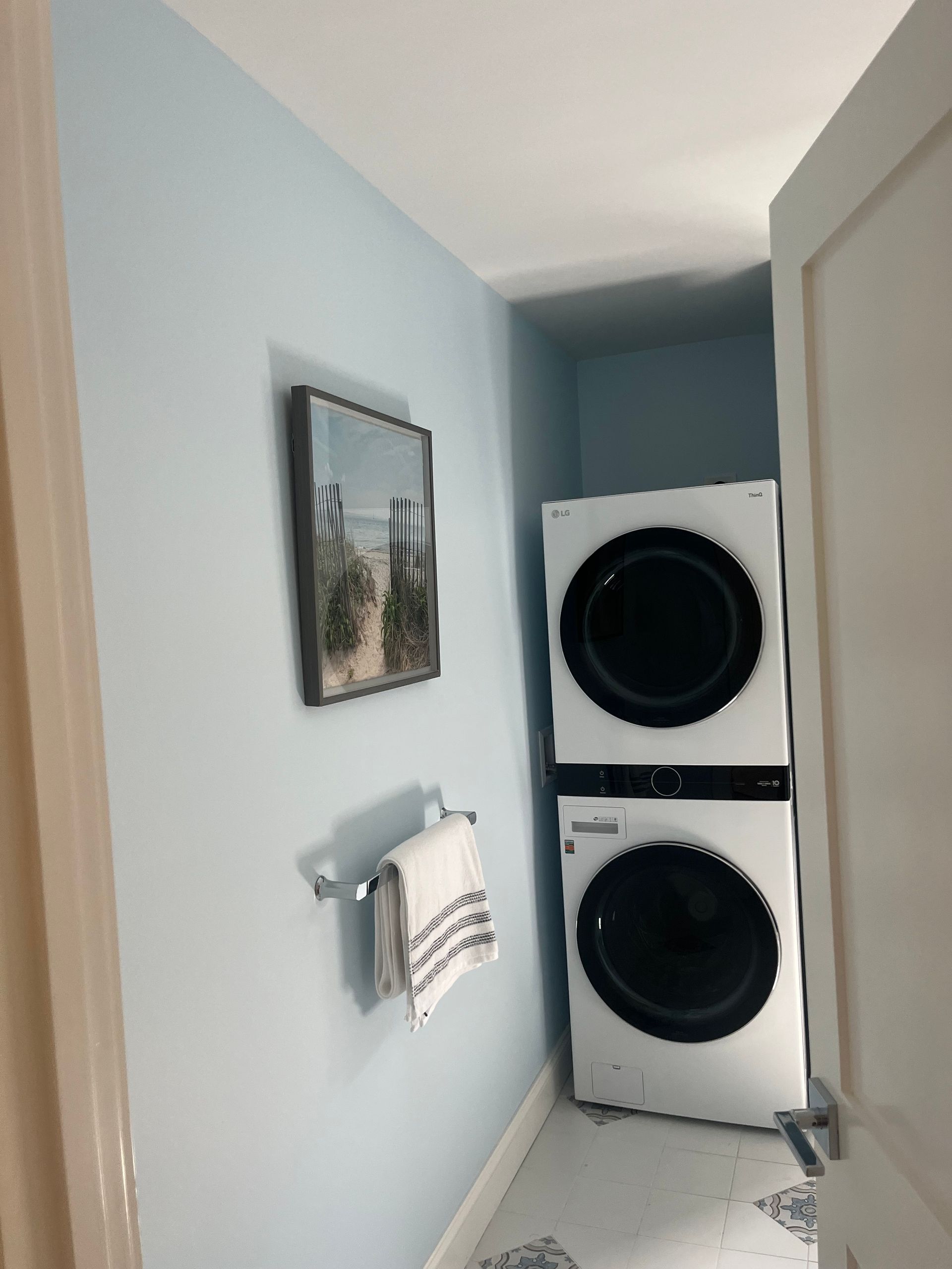 Stackable washer and dryer in a small, pale blue laundry room with artwork and towel rack.