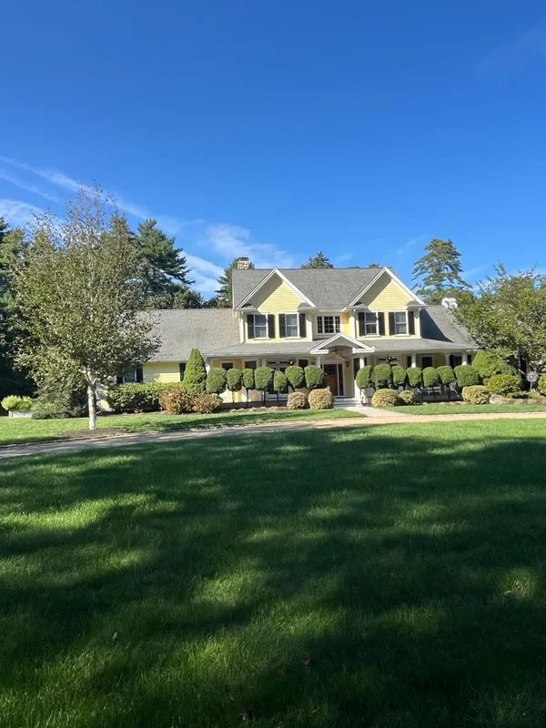 Yellow house with black shutters and manicured shrubs on a green lawn under a blue sky.