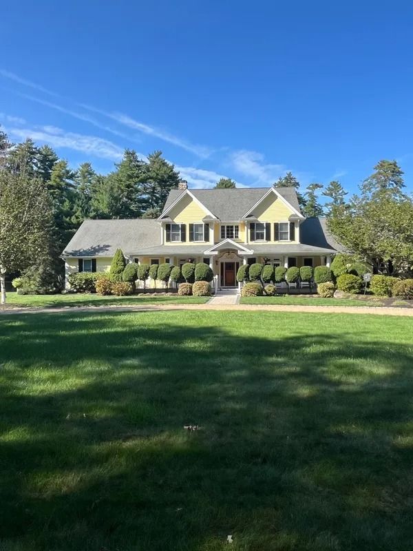 Yellow house with black shutters, surrounded by green lawn and trees under a blue sky.