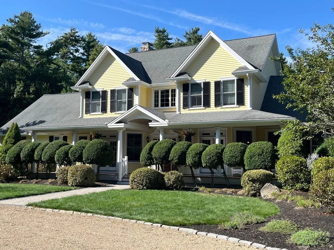 Yellow house with black shutters, green bushes, and a gray roof on a sunny day.