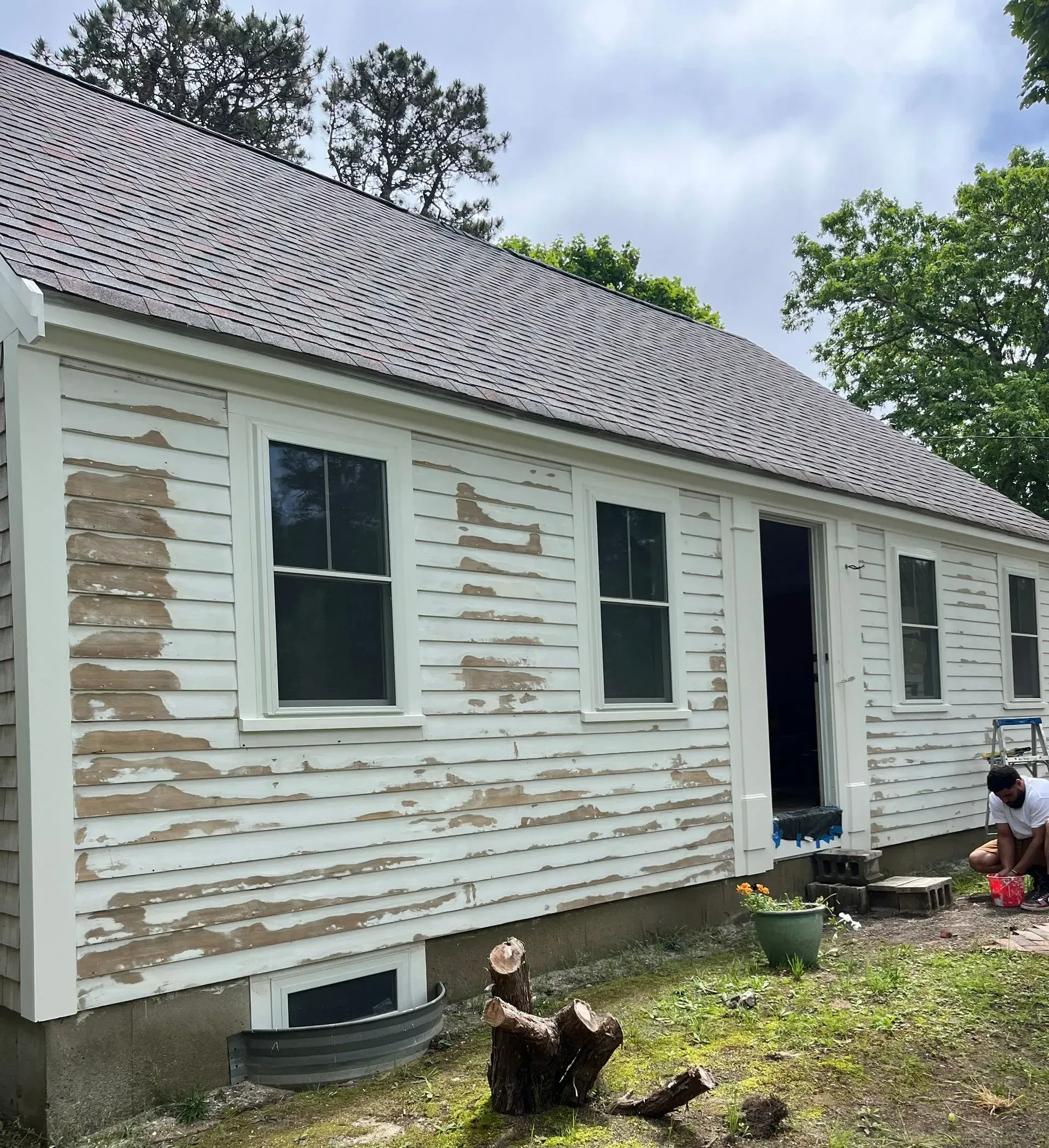 White house with peeling paint, windows, and an open door. Person outside, working on the side of the house.