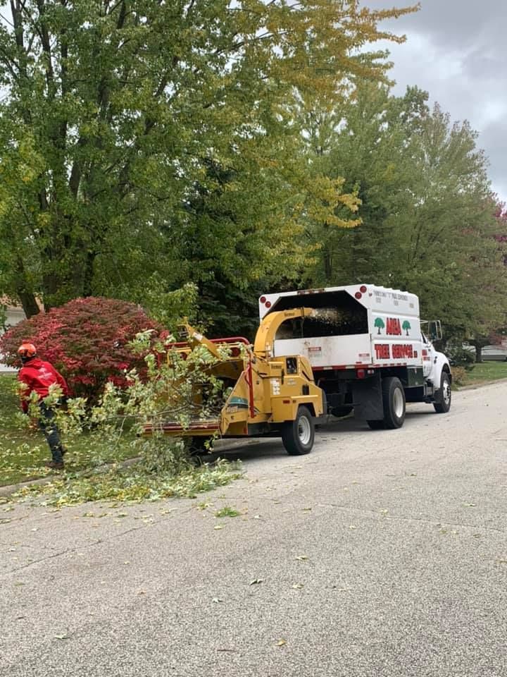 A worker in a red uniform feeds tree branches into a yellow wood chipper attached to a truck on a residential street.