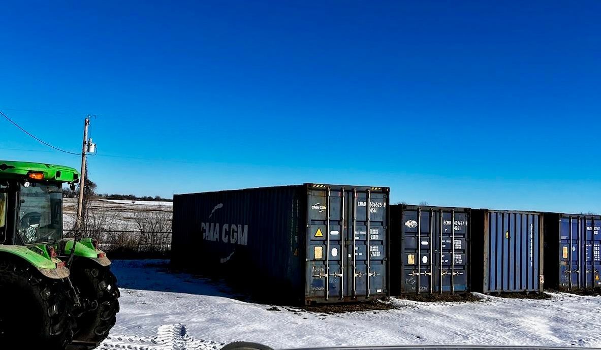 A green tractor is parked next to a row of shipping containers in the snow