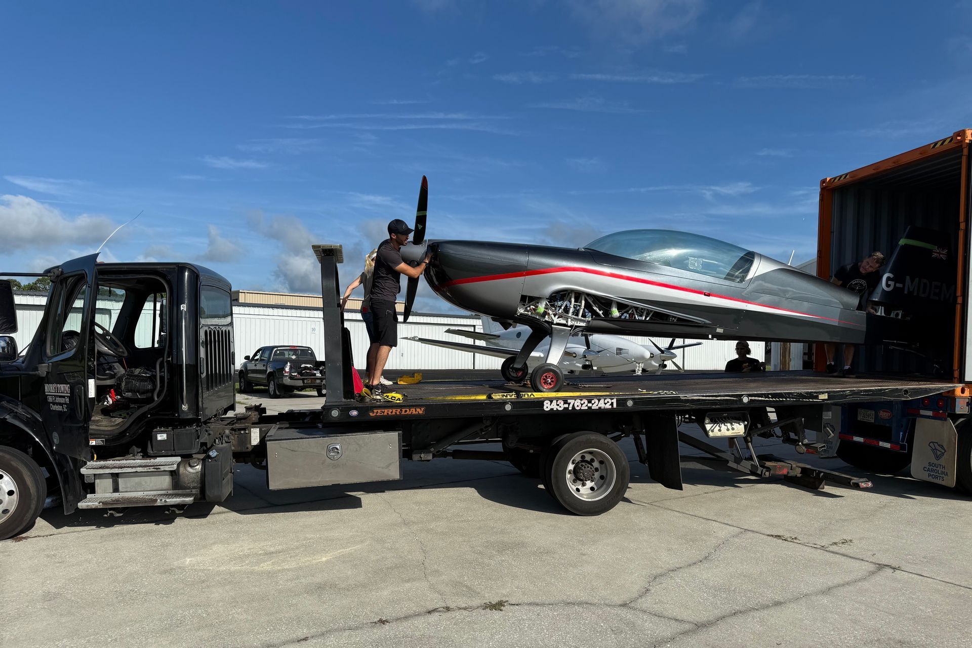 A small airplane being loaded onto a flatbed truck from a shipping container on a sunny day. A person is assisting.