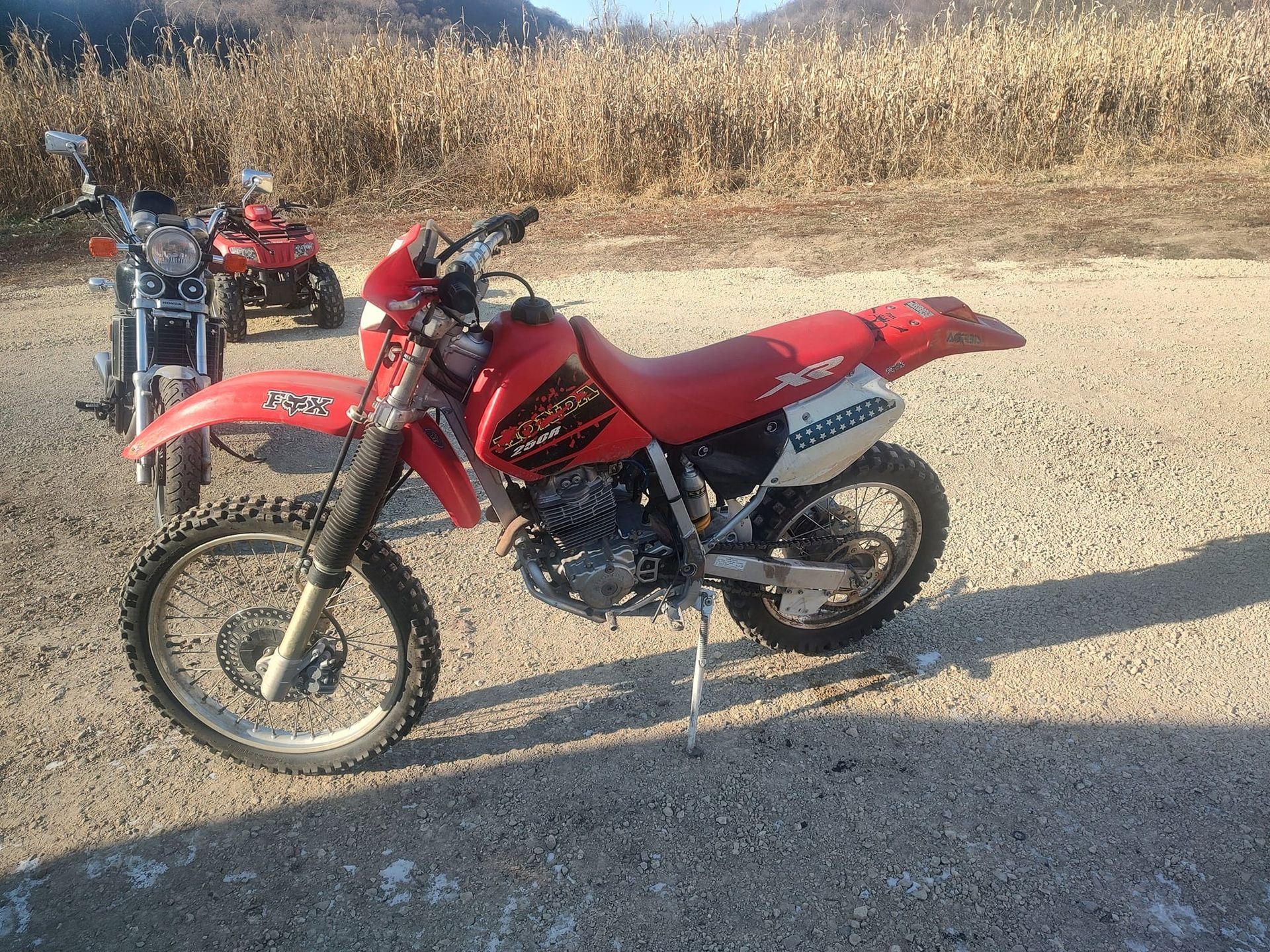 Red Honda XR650L motorcycle parked on a gravel road, other motorcycles in the background, field in sunlight.