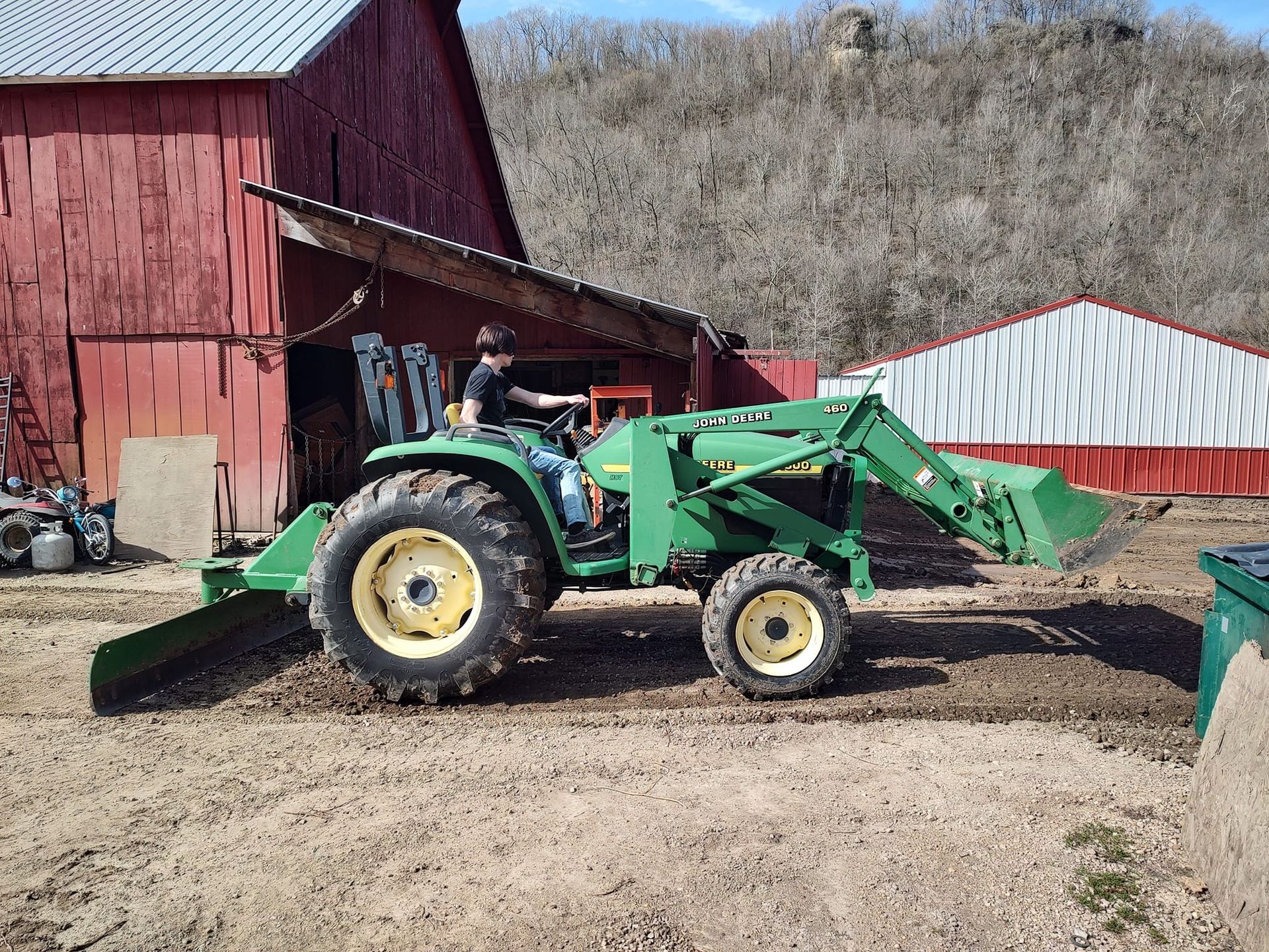 A person operating a green John Deere tractor with a front loader near a red barn.