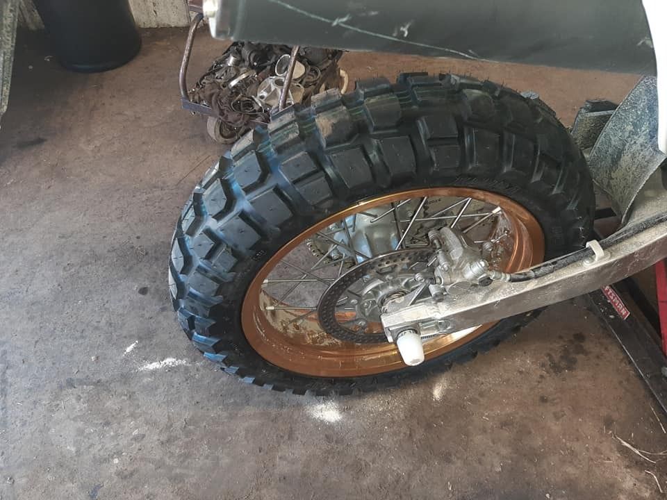 Motorcycle rear wheel with a knobby tire, gold rim, and silver swingarm.