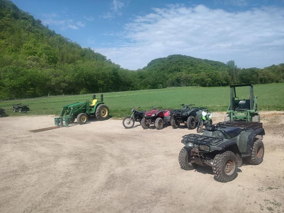 Green tractor and ATVs parked on a dirt lot in front of a hillside on a sunny day.