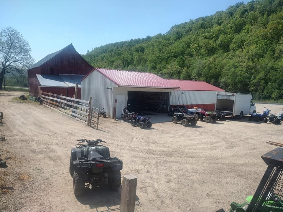 A barn with ATVs parked in front, dirt driveway, and a tree-covered hillside in the background.
